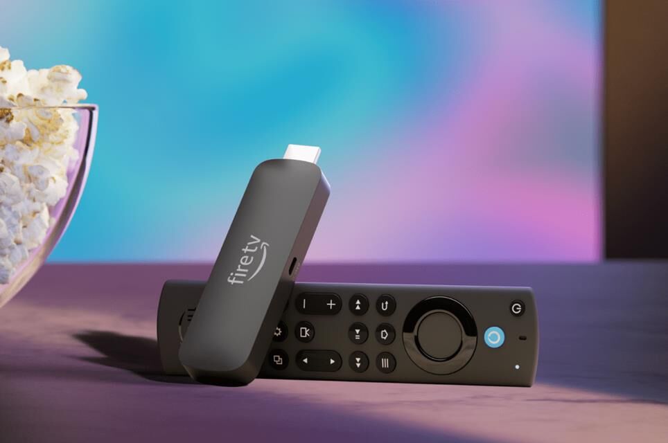 Thiết bị phát trực tuyến Amazon Fire TV Stick 4K Max cùng điều khiển từ xa đặt cạnh một bát bỏng ngô trước TV.