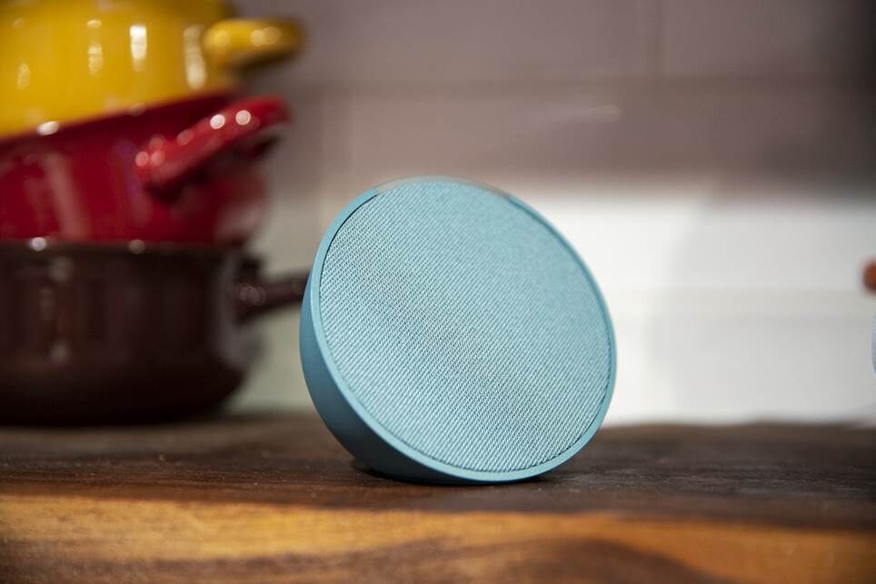Teal Amazon Echo Pop nhìn từ một góc với những chậu cây nhiều màu sắc ở phía sau.