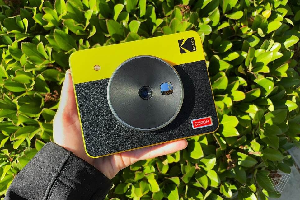 Chiếc máy ảnh Kodak Mini Shot 3 Retro màu vàng và đen được giơ ngang với một bàn tay và một bụi cây làm nền.